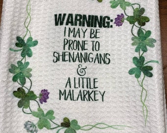 Puede incluir: Toalla de cocina blanca con el texto "WARNING: I MAY BE PRONE TO SHENANIGANS & A LITTLE MALARKEY" en verde oscuro. La toalla está decorada con tréboles verdes y flores moradas.