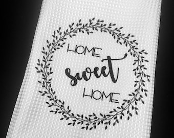 Puede incluir: Toalla de cocina blanca con tejido de gofre texturizado. Una corona negra rodea las palabras "HOME sweet HOME" en una fuente cursiva. La toalla está doblada y se muestra sobre un fondo negro.