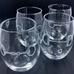 May include: Four clear stemless wine glasses with a horse bit design etched on the side. The glasses are transparent, allowing the contents to be visible. The horse bit design is a light color, contrasting against the clear glass. The glasses are arranged on a dark surface.