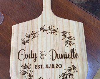 Puede incluir: Una pala de pizza de madera clara con un mango y un agujero circular. La pala está grabada con los nombres "Cody & Danielle" y la fecha "EST. 4.18.20" rodeados por un diseño de corona floral.