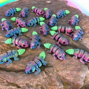May include: A collection of 17 small, segmented, plastic lizards with iridescent purple, blue, and green paint. The lizards have green tails and are arranged in a circle on a brown rock.