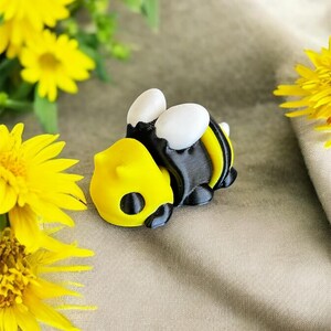 May include: A 3D printed black and yellow bee figurine with white wings. The bee is lying on its side with its head facing the left. The bee is on a light brown fabric background.