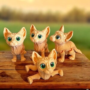 May include: Four tan-colored Sphynx cat figurines with large green eyes are arranged on a wooden surface. The cats are in various poses, some standing and others with their legs outstretched. The background is blurred, suggesting an outdoor setting.
