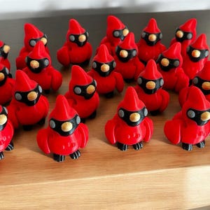 May include: A collection of red cardinal figurines with black masks and beaks, and gray accents. The figurines are arranged on a wooden surface. The cardinals are a vibrant red color and have a cartoonish design.