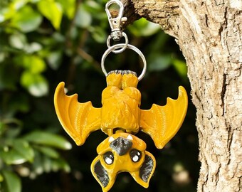 May include: A yellow bat-shaped keychain with black accents, hanging upside down. The bat has large, expressive eyes and detailed wings. The keychain is attached to a silver clasp and ring, perfect for keys or bags.
