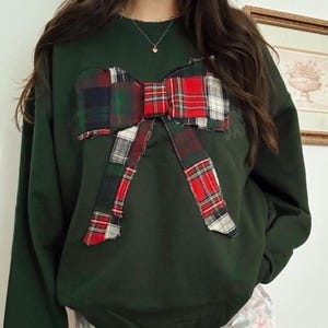 Puede incluir: Una sudadera verde oscuro con un gran lazo a cuadros en rojo, verde y blanco. La modelo también lleva unos pantalones cortos blancos con un ribete rosa con volantes. Se ve un collar de oro.
