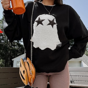 May include: Black sweatshirt with a white ghost design and star eyes. The person is wearing pink and white striped trousers and carrying a pumpkin-shaped purse with a bat. A mug of orange liquid is held in the hand.