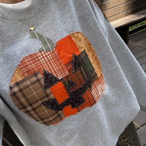 Peut inclure: Sweat-shirt gris avec un motif citrouille en patchwork. La citrouille est composée de divers motifs de tissu orange, marron, vert et noir. Le sweat-shirt a un col rond et des manches longues, parfait pour l'automne.
