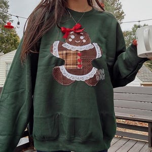 May include: A dark green sweatshirt featuring a gingerbread man applique with a red bow. The gingerbread man has a brown body, plaid torso, and white lace trim. The text "FUN DAY" is on the lower right. A white mug with floral designs is held in the hand.