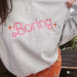 May include: A light gray sweatshirt with the word "Boring" embroidered in pink script across the chest. The word is flanked by a pink and orange starburst on the left and a green and orange starburst on the right. The sweatshirt is part of a casual outfit.