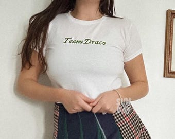 Może przedstawiać: Biała koszulka z napisem "Team Draco" w zielonych literach. Osoba nosi spódnicę w kratę w kolorze niebieskim, zielonym i czerwonym.