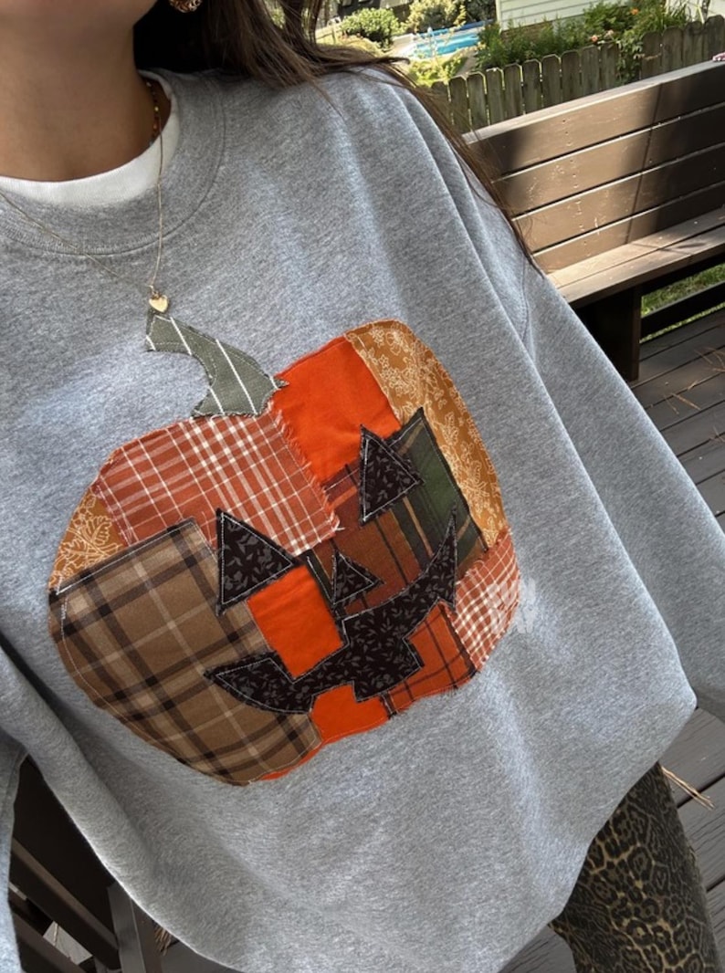 K&ouml;nnte beinhalten: Ein graues Sweatshirt mit Rundhalsausschnitt und einer gro&szlig;en, mittigen K&uuml;rbis-Applikation aus verschiedenen Stoffst&uuml;cken in Orange, Braun und Gr&uuml;n mit Karomustern, Blumenmustern und unifarbenen Stoffen sowie schwarzen Ausschnitten.