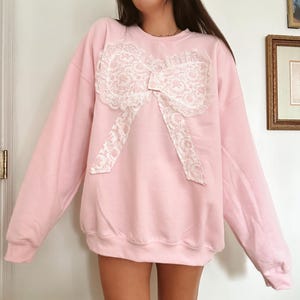 May include: A light pink sweatshirt featuring a large, white lace bow appliqué on the front. The bow has a floral pattern and is centered on the chest. The sweatshirt has long sleeves and a crew neck.