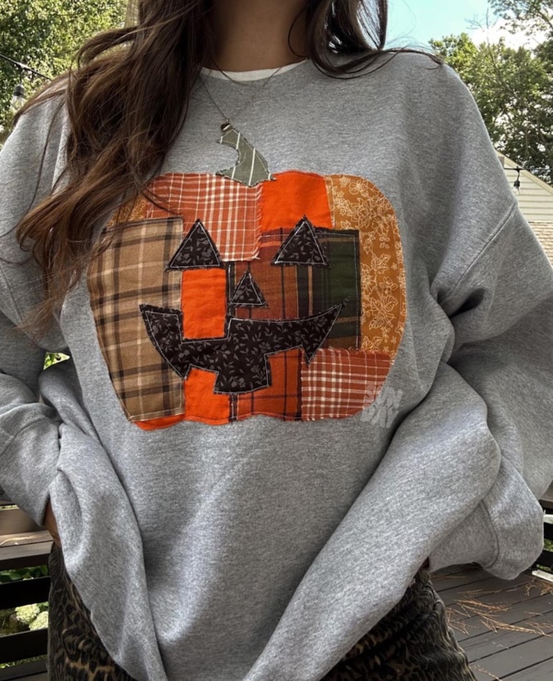 K&ouml;nnte beinhalten: Ein graumeliertes Sweatshirt mit Rundhalsausschnitt und einer gro&szlig;en, zentralen K&uuml;rbis-Applikation aus verschiedenen Stoffst&uuml;cken in Orange, Braun und Gr&uuml;n mit Karomustern, Blumenmustern und unifarbenen Stoffen sowie einem dunklen Gesicht.