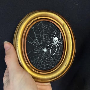 Peut inclure: Cadre ovale doré avec une œuvre d'art représentant une araignée blanche à tête de mort sur une toile d'araignée, sur fond noir. Le cadre est doré avec un bord intérieur cuivré. L'œuvre est tenue dans une main.