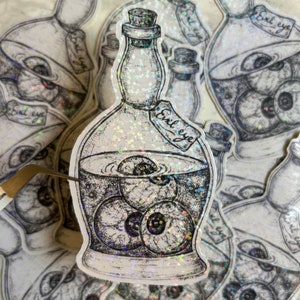 May include: A sticker of a glass bottle filled with eyeballs. The bottle has a label that reads "Evil eye".