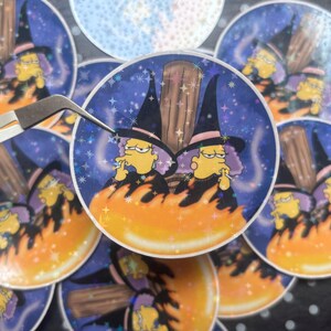 May include: Circular stickers with a cartoon witch character, yellow skin, purple hair, and a black hat. The witch is smoking, set against a starry night sky and flames. A pair of tweezers holds one sticker.