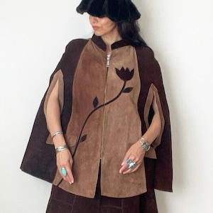 May include: A vintage suede ensemble in shades of brown. The outfit includes a cape with a floral design, a skirt, and knee-high boots. A black fur hat completes the look. The cape features a zipper closure and a unique cutout design for the arms.