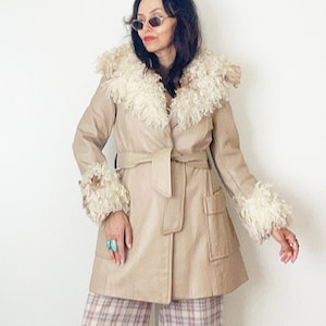 May include: A beige coat featuring a large cream-colored curly faux fur collar and matching cuffs, a belted waist, and two front pockets. The coat is paired with wide-leg trousers in a light purple, white, and beige plaid pattern and brown shoes.