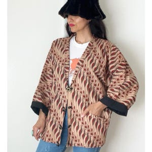 May include: A patterned jacket with a beige background and red and brown designs. The jacket has black trim on the sleeves and pockets. The person in the image is wearing a black fur hat, a white t-shirt, and blue jeans.