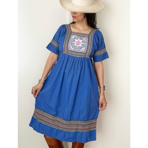 May include: A blue dress with short sleeves and a square neckline. The dress features a colorful embroidered design on the bodice and hem. The person is wearing a cream-colored hat and brown boots.