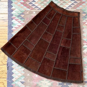 May include: A brown patchwork skirt with a flared silhouette. The skirt is constructed from rectangular and square pieces of leather, stitched together with dark brown thread. The hemline and waistband are also trimmed with the same dark brown thread.