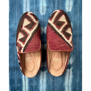 May include: A pair of slip-on mules featuring a woven textile upper with a geometric pattern in shades of deep red, dark brown, and cream. The shoes have a smooth tan leather trim along the opening and a tan leather insole with visible text.