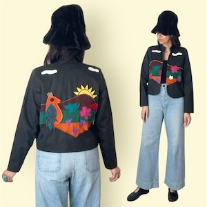 May include: Black jacket with colorful embroidered landscape design featuring a bird, sun, and flowers. The jacket has long sleeves and a round collar. The model is wearing light blue jeans and a black hat.