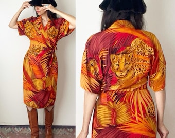 May include: A wrap dress with a vibrant tropical print featuring leopards and palm leaves in shades of orange, red, and yellow. The dress has short sleeves and a tie waist. The model is wearing brown boots and a black hat.