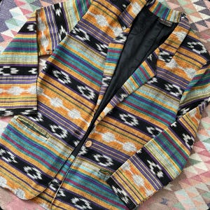 May include: A colorful blazer with a geometric pattern in shades of orange, purple, teal, and black. The blazer has a single button closure and two front pockets. The interior lining is black.