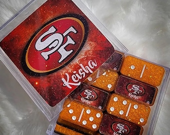 May include: A clear acrylic box containing a set of dominoes. The box lid features the San Francisco 49ers logo and the name "Keisha". The dominoes are orange and burgundy with white dots.