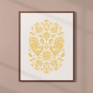 A framed artwork with a symmetrical design featuring two roosters and flowers in yellow on a white background. The wooden frame hangs elegantly on a light brown wall.