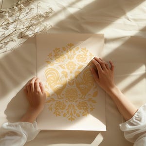 Two hands hold a art print with an intricate yellow design, including floral elements and two symmetrically opposing roosters. The paper lies on a cream-colored fabric, surrounded by shadows and dried flowers.