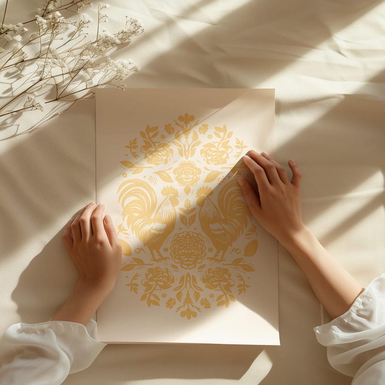 Two hands hold a art print with an intricate yellow design, including floral elements and two symmetrically opposing roosters. The paper lies on a cream-colored fabric, surrounded by shadows and dried flowers.