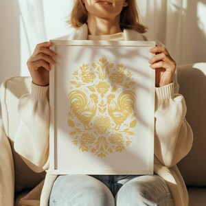 A person in a white sweater is seated on a light-colored couch, holding a framed artwork featuring a symmetrical design with yellow floral patterns and two birds facing each other. Sunlight illuminates the scene as the perfect birthday gift.