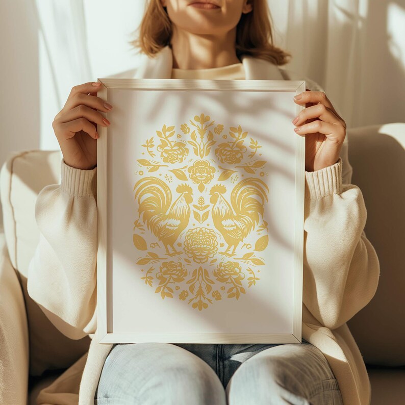 A person in a white sweater is seated on a light-colored couch, holding a framed artwork featuring a symmetrical design with yellow floral patterns and two birds facing each other. Sunlight illuminates the scene as the perfect birthday gift.