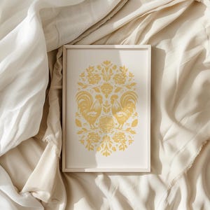 A framed artwork featuring two stylized golden birds and intricate floral patterns is placed on a soft, cream-colored fabric background with gentle folds and light streaming in from one side, resembling the elegance of vintage clothes.