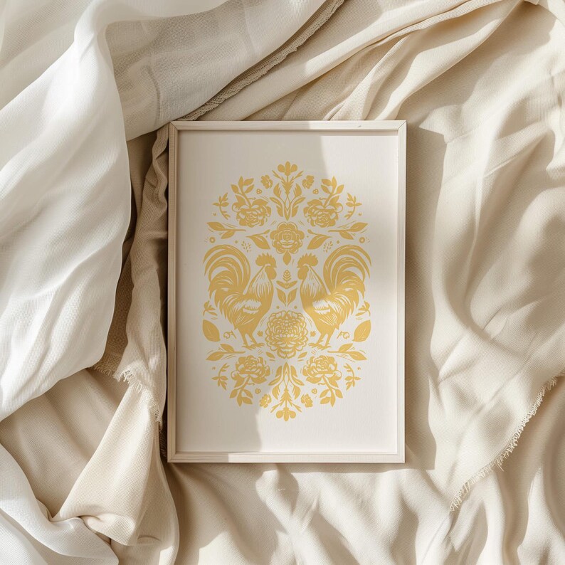 A framed artwork featuring two stylized golden birds and intricate floral patterns is placed on a soft, cream-colored fabric background with gentle folds and light streaming in from one side, resembling the elegance of vintage clothes.