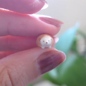 May include: A miniature ceramic hedgehog figurine. The hedgehog has a brown body with white spines and a white face. It has small, dark eyes and is held between fingers.