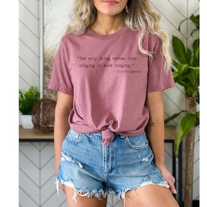 Puede incluir: Una camiseta color malva con una cita de Ella Fitzgerald: "Lo único que es mejor que cantar es cantar más."