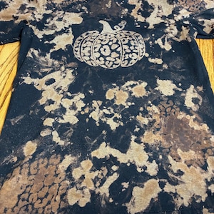 May include: A black short-sleeve t-shirt featuring a bleached tie-dye effect with irregular tan and brown patches. The center of the shirt displays a graphic of a pumpkin filled with a leopard print pattern in shades of tan and dark brown.