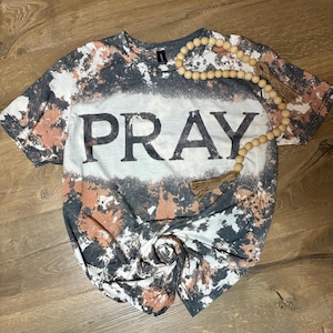 May include: A short-sleeve t-shirt with a bleached, mottled pattern in shades of charcoal gray, white, and muted orange. The center features the word PRAY in large, bold, black capital letters. A string of light-colored wooden beads with a tassel is included.