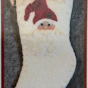 Könnte beinhalten: Ein weißer Weihnachtsstrumpf mit einem Weihnachtsmann-Gesicht. Der Weihnachtsmann hat eine rote Mütze mit einem grünen Stechpalmenblatt. Der Strumpf besteht aus einem weichen, flauschigen Material.