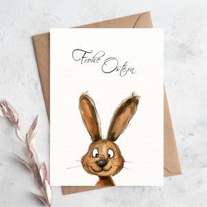 Pu&ograve; includere: Un biglietto bianco con il testo "Frohe Ostern" in corsivo nero. Un coniglio di cartone animato con pelliccia marrone spunta dalla parte inferiore del biglietto.