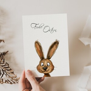 Pu&ograve; includere: Un biglietto bianco con il testo "Frohe Ostern" in calligrafia nera. Un coniglio dei cartoni animati con la pelliccia marrone e gli occhi grandi sta sbirciando dal fondo del biglietto.