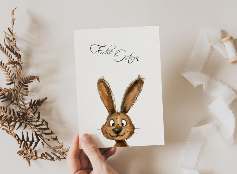Pu&ograve; includere: Un biglietto bianco con il testo "Frohe Ostern" in calligrafia nera. Un coniglio dei cartoni animati con la pelliccia marrone e gli occhi grandi sta sbirciando dal fondo del biglietto.
