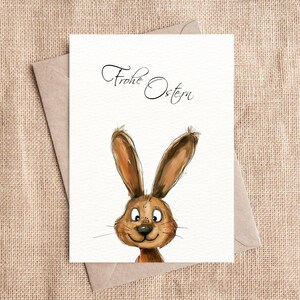 Pu&ograve; includere: Un biglietto bianco con il testo "Frohe Ostern" scritto in corsivo nero. Un coniglio dei cartoni animati con la pelliccia marrone e gli occhi grandi spunta dal fondo del biglietto.