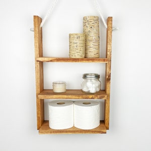 May include: A rustic wooden shelf with three tiers, hanging from a white rope. The shelf is filled with decorative items, including candles, a jar of cotton balls, and two rolls of toilet paper.