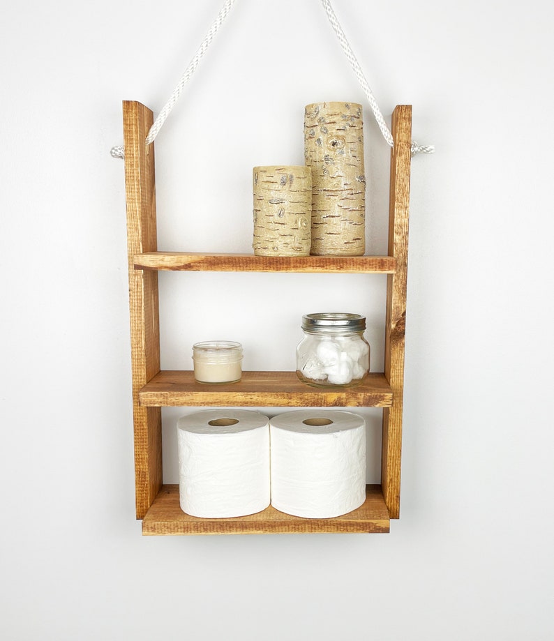 May include: A rustic wooden shelf with three tiers, hanging from a white rope. The shelf is filled with decorative items, including candles, a jar of cotton balls, and two rolls of toilet paper.