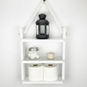 May include: White wooden shelf with two shelves, hanging from a hook on a white wall. The top shelf has a black lantern with a white star design. The middle shelf has a clear glass jar with white cotton balls and a small white candle. The bottom shelf has two rolls of white toilet paper.